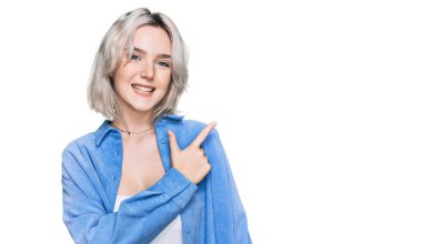 Yüzünde mutlu ve doğal bir ifadeyle elleri ve parmaklarıyla işaret eden güler yüzlü, gündelik kıyafetler giyen genç sarışın kız. 