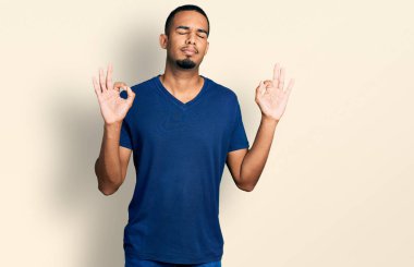 Rahat bir tişört giyen, gözleri kapalı gülümseyen genç Afro-Amerikan erkeği parmaklarıyla meditasyon hareketi yapıyor. yoga konsepti. 