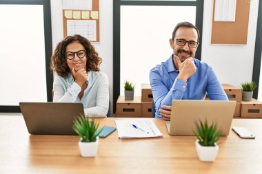 Orta yaşlı İspanyol bir kadın ve ofiste dizüstü bilgisayarı olan bir adam kameraya kendinden emin bir şekilde bakıyor kollarını kavuşturmuş ve çenesini kaldırmış gülümsüyor. Olumlu düşünmek. 