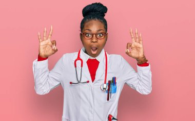 Doktor üniforması ve steteskop giyen genç Afro-Amerikan kadın parmaklarıyla onay sembolü yaparken şaşırmış ve şok olmuş görünüyor. çılgın ifade 