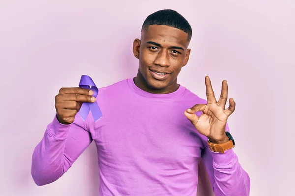 Young black man holding purple ribbon awareness doing ok sign with fingers, smiling friendly gesturing excellent symbol
