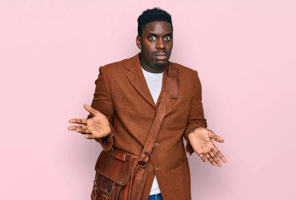Handsome business black man wearing leather bag clueless and confused expression with arms and hands raised. doubt concept.