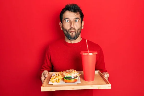 Handsome man with beard eating a tasty classic burger with fries and soda making fish face with mouth and squinting eyes, crazy and comical.