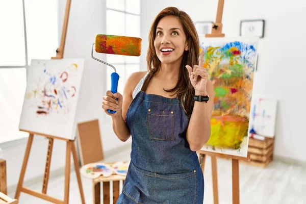 若いラテンアーティストの女性を保持しているペイントローラースタンドでアートスタジオ驚きでアイデアや質問を指差す指で幸せな顔 ナンバーワン — ストック写真