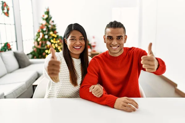 一对年轻的拉丁夫妇坐在桌上 坐在圣诞树旁 赞成用手做积极的手势 微笑着竖起大拇指 为成功感到高兴 优胜手势 — 图库照片