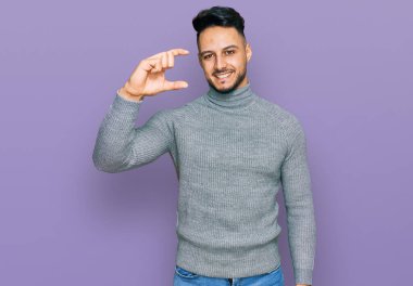 Gündelik kıyafetler giyen, gülümseyen, kendine güvenen, el hareketleriyle hareket eden, parmaklarıyla işaret yapan ve kameraya bakan genç bir Arap. ölçüm kavramı. 