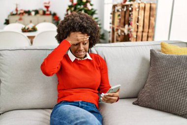 Afro saçlı, akıllı telefon kullanan genç Afro-Amerikalı kadın Noel ağacının kenarında oturuyor ve elleri kafasında, şaşırmış ve kızgın yüzüyle stresli. 