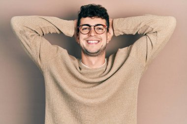 Genç İspanyol adam günlük kıyafetler giyiyor ve gözlük takıyor rahatlıyor ve esniyor, kolları ve elleri başının ve boynunun arkasında mutlu bir şekilde gülümsüyor. 