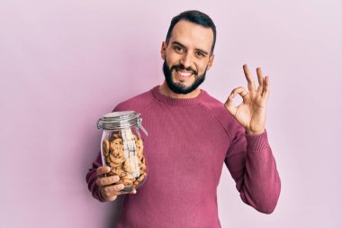 Sakallı genç adam elinde bir kavanoz çikolatalı kurabiye tutuyor. Parmaklarıyla imza atıyor, gülümseyerek mükemmel bir sembol oluşturuyor. 