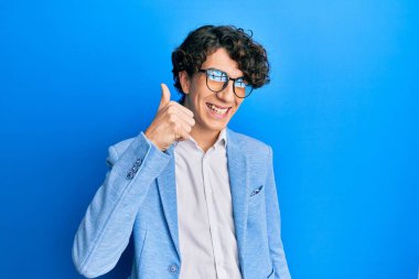 İş ceketi ve gözlük takan İspanyol genç adam mutlu ve pozitif gülümsüyor, başparmağıyla mükemmel ve onay işareti yapıyor. 