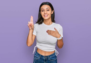 Latin asıllı genç bir kız, günlük beyaz tişört giyiyor, gülümsüyor, elleri göğsünde ve parmakları havada küfür ediyor, sadakat yemini ediyor. 