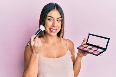 Makyaj fırçası tutan ve yüzünde mutlu ve havalı bir gülümsemeyle gülümseyen genç İspanyol kadın. diş gösterme. 