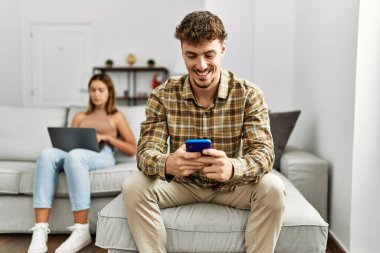 Genç İspanyol çift dizüstü bilgisayar ve akıllı telefon kullanarak mutlu bir şekilde gülümsüyor. Evdeki kanepede oturuyor..