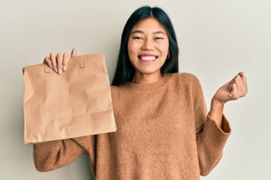 Elinde kağıt torba tutan genç Çinli kadın gururla bağırıyor, zaferi kutluyor ve kollarını kaldırarak heyecanlanıyor. 