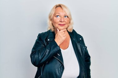 Orta yaşlı, deri ceket giyen, elleri çenesinde soru soran, düşünceli bir kadın. Düşünceli bir yüzle gülümsüyor. şüphe kavramı. 