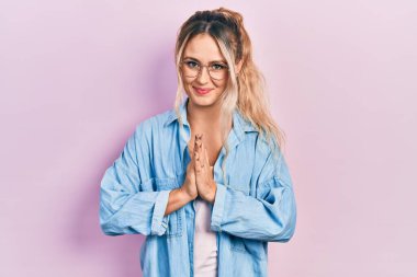 Güzel, genç, sarışın bir kadın. Günlük kıyafetler ve gözlük takıyor. Elleriyle dua ediyor, af diliyor.. 