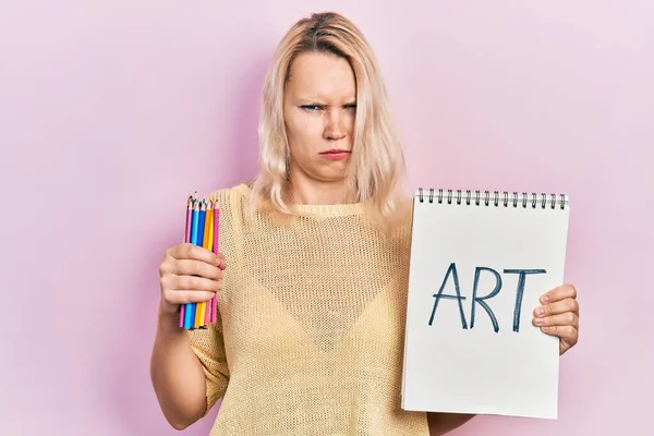 美しい白人ブロンドの女性は アートノートと色鉛筆懐疑的で緊張し 問題のために動揺して眉をひそめています 否定的な人 — ストック写真