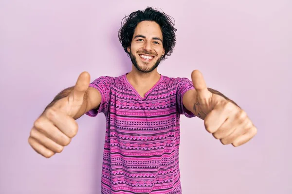 手で積極的なジェスチャーを行うことを承認カジュアルなピンクのTシャツを着てハンサムなヒスパニック系の男性 親指アップ笑顔と成功のために幸せ 勝者のジェスチャー — ストック写真