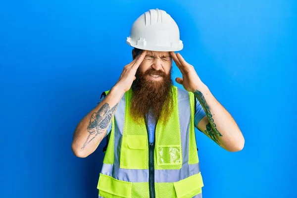 Redhead man with long beard wearing safety helmet and reflective jacket with hand on head for pain in head because stress. suffering migraine.