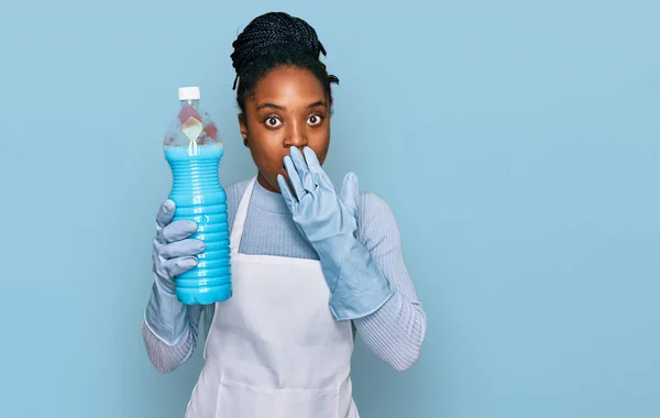 Mujer Afroamericana Joven Con Delantal Sosteniendo Botella Detergente Cubriendo Boca —  Fotos de Stock