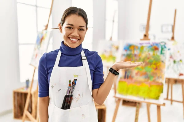 Young brunette woman at art studio smiling cheerful presenting and pointing with palm of hand looking at the camera.