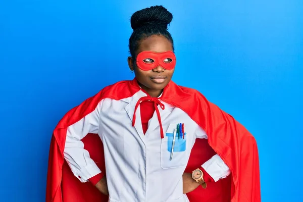 身穿医生制服和超级英雄服装的年轻非洲裔美国女人面带微笑地看着旁边 目不转睛地思考着 — 图库照片