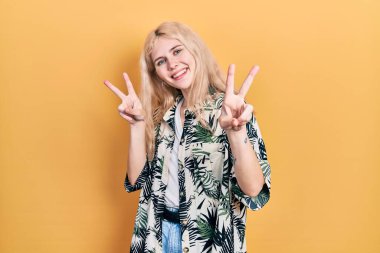 Sarı saçlı, tropikal tişörtlü güzel beyaz kadın kameraya bakıp zafer işareti yapıyor. İki numara.. 