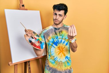 İspanyol asıllı genç adam paletle resim çiziyor. Resim sehpası ayakta duruyor. El ve parmaklarıyla italyan hareketi yapıyor. 