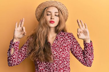 Güzel sarışın beyaz kadın yaz şapkası takıyor rahatlıyor ve gözleri kapalı gülümsüyor parmaklarıyla meditasyon hareketi yapıyor. yoga konsepti. 