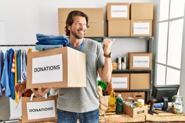 Gönüllü olarak bağış kutusunu tutan yakışıklı orta yaşlı adam arka tarafı işaret ediyor. El ve başparmakları havada, kendinden emin gülümsüyor. 