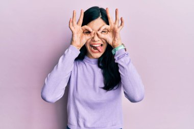 Günlük kıyafetler giyen genç İspanyol kadın dürbün gibi jest yapıyor, gözleri parmaklarından bakıyor. çılgın ifade. 