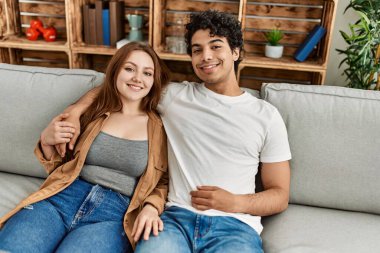 Mutlu gülümseyen ve sarılan genç çift evdeki kanepede oturuyor..