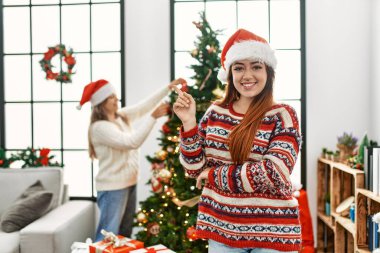 Evdeki güzel çift Noel ağacının yanında durup gülümsüyor mutlu bir şekilde elleri ve parmakları yana doğru işaret ediyor. 