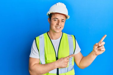 Güvenlik kaskı takan yakışıklı genç bir adam ve yansıtıcı ceket giymiş gülümseyen ve kameraya bakan iki eliyle ve parmaklarıyla yan tarafı gösteren.. 