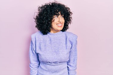 Orta Doğulu genç bir kadın gündelik kıyafetler giyip yüzünde gülümseme, doğal bir ifade ile yan yana bakıyor. Kendinden emin gülen. 