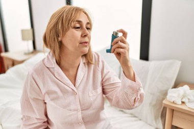 Orta yaşlı bir kadın astım ilacı kullanıyor. Evdeki yatakta oturuyor..