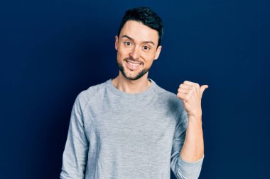 Gündelik kıyafetler giyen, gülümseyen, gülümseyen ve başparmağıyla yan tarafı gösteren İspanyol genç adam.. 