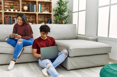 Evde dizüstü bilgisayar ve akıllı telefon kullanarak mutlu gülümseyen genç Latin çift..