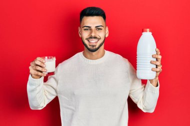Sakallı, elinde bir bardak süt ve plastik şişeyle gülümseyen genç, İspanyol bir adam. diş gösterme. 