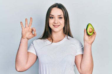 Ortadaki avokadoyu tutan genç İspanyol kız parmaklarıyla imza atıyor, gülümseyerek mükemmel bir sembol oluşturuyor. 