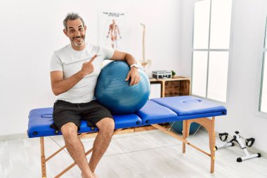 Orta yaşlı İspanyol bir adam ağrı kesici kliniğinde elinde neşeli bir pilates topu tutarken yüzünde bir gülümseme, mutlu ve doğal bir ifadeyle elini ve parmağını yana kaldırıyor. 