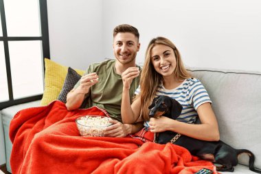 Genç İspanyol çift gülümsüyor. Mutlu bir şekilde kanepede oturmuş, evde köpekleriyle film izliyorlar..