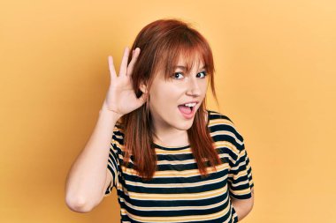 Sıradan çizgili tişört giyen, kulağı kulağında gülümseyen ve dedikoduları ve dedikoduları duyan kızıl saçlı genç bir kadın. Sağırlık kavramı. 