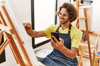 Genç İspanyol ressam gülümsüyor mutlu çizimler yapıyor ve resim stüdyosunda akıllı telefon kullanıyor..