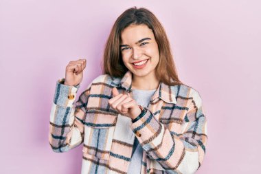 Gündelik giysiler giyen genç beyaz kız arka tarafı işaret ediyor. Elleri havada, kendinden emin bir şekilde gülümsüyor. 