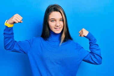 Balıkçı yaka kazak giyen genç esmer kız gururla gülümseyen kol kaslarını gösteriyor. fitness konsepti. 