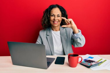Ofiste çalışan orta yaşlı güzel bir kadın bir fincan kahve içiyor gülümsüyor ve kendinden emin el hareketleriyle küçük beden işareti yapıyor parmaklarıyla ve kamerayla bakıyor. ölçüm kavramı. 