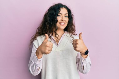 Kıvırcık saçlı, sıradan giyinen genç esmer kadın. Başarı işareti yapıyor. Elleriyle olumlu jest yapıyor. Neşeli ifade ve kazanan jest. 