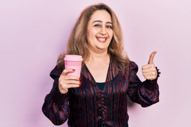 Orta yaşlı, beyaz bir kadın kahvesini yudumluyor mutlu ve pozitif gülümsüyor, başparmağıyla mükemmel ve onay işareti yapıyor. 