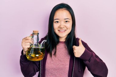 Zeytin yağı tutan genç Çinli kız mutlu ve pozitif gülümsüyor, başparmağıyla mükemmel ve onay işareti yapıyor. 